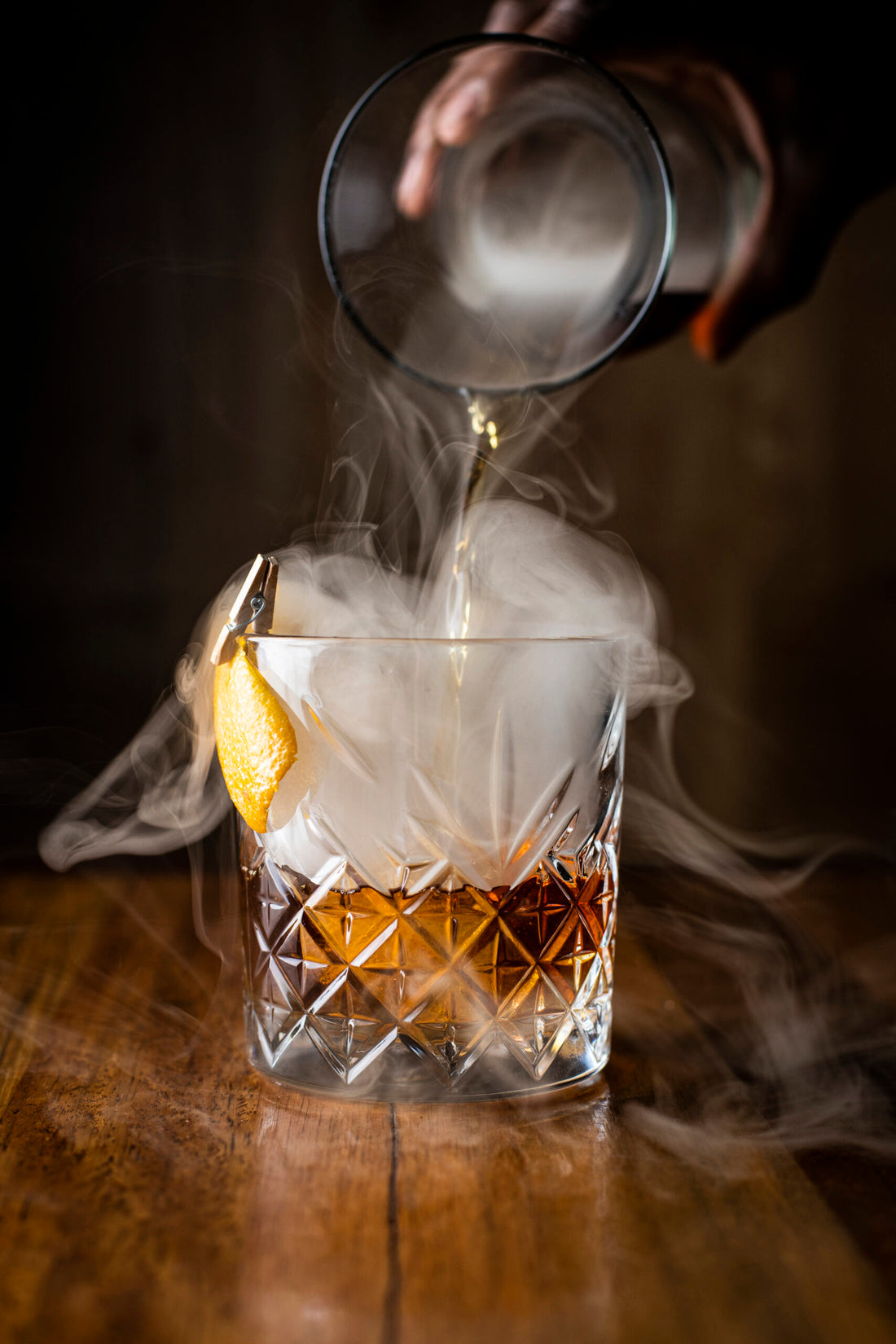 An impressive photo of a glass of whiskey being poured by hand. There is smoke billowing out from the decanter into the glass. The glass is garnished with an orange peel.