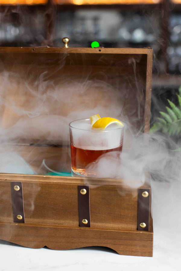 A deep amber cocktail in a rocks glass with a large ice cube and lemon twist garnish, presented inside an open wooden chest box surrounded by billowing smoke