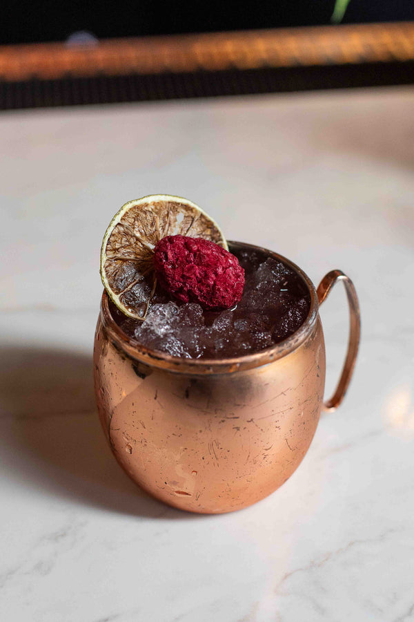 A dark berry cocktail in a copper mule mug filled with crushed ice, garnished with a dried lime wheel and a freeze-dried raspberry, on a white marble surface
