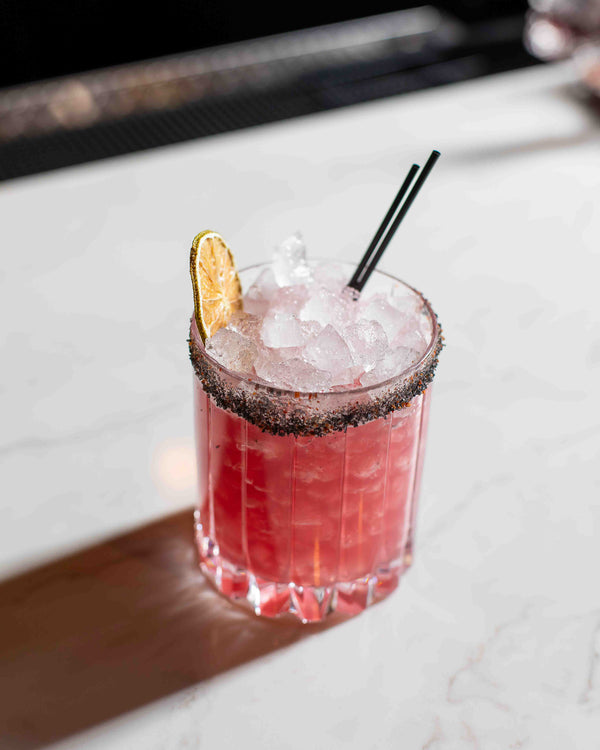 A deep pink cocktail in a ribbed rocks glass with a dark spiced salt rim, filled with crushed ice and garnished with a dried lime wheel and two black straws, on a white marble surface