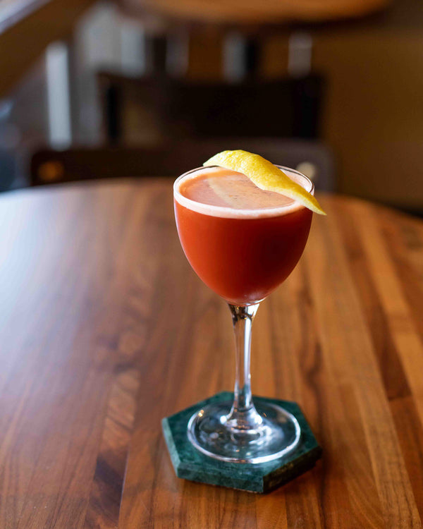 A deep red cocktail in a stemmed wine-style glass with a yellow lemon twist garnish, served on a green marble hexagon coaster on a warm wood bar top
