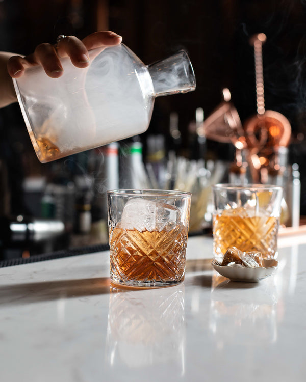 A bartender pouring smoke from a glass decanter over two amber old fashioned cocktails with large clear ice cubes in cut crystal rocks glasses, set on a white marble bar with copper bar tools in the background