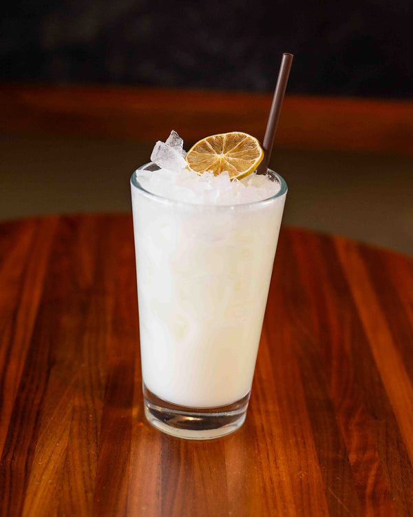 A creamy white cocktail in a tall pint glass, filled with crushed ice and garnished with a dried lime wheel and a brown paper straw, set on a warm wood bar top