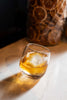 Glass of whiskey with ice cubes on a marble surface, with a wooden barrel in the background.