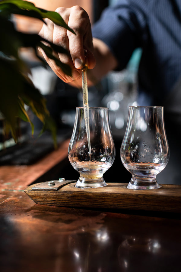 A hand adding drops from a glass dropper into a Glencairn tasting glass, one of two glasses on a narrow wooden flight board, representing the spirit enrichment experience at Northwest Spirits