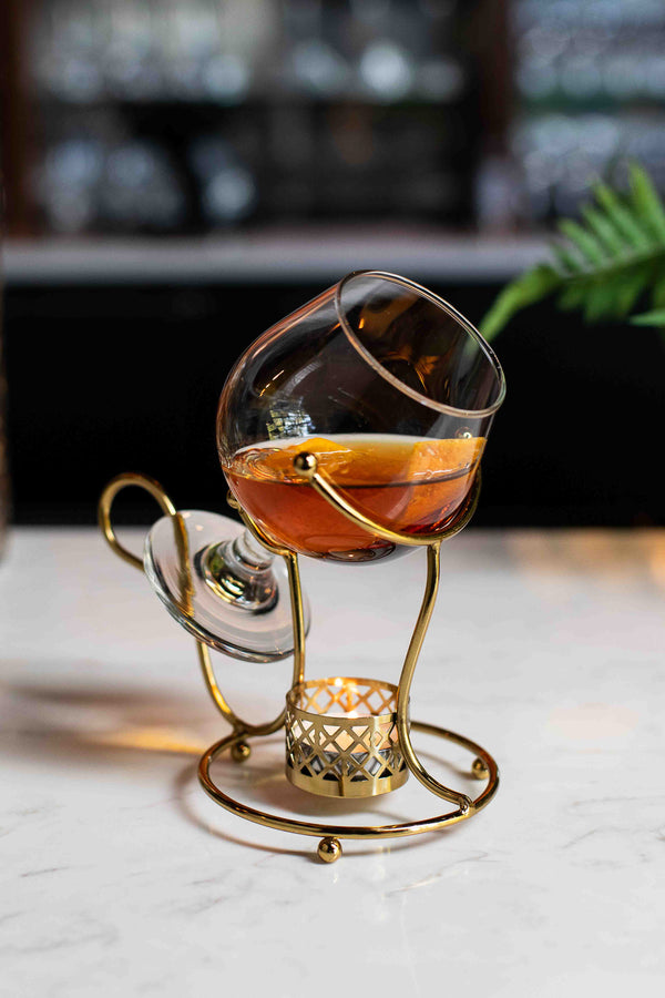 A rich amber cocktail in a tilted brandy glass balanced on an ornate gold metal stand with a small candle warmer beneath, served on a white marble surface