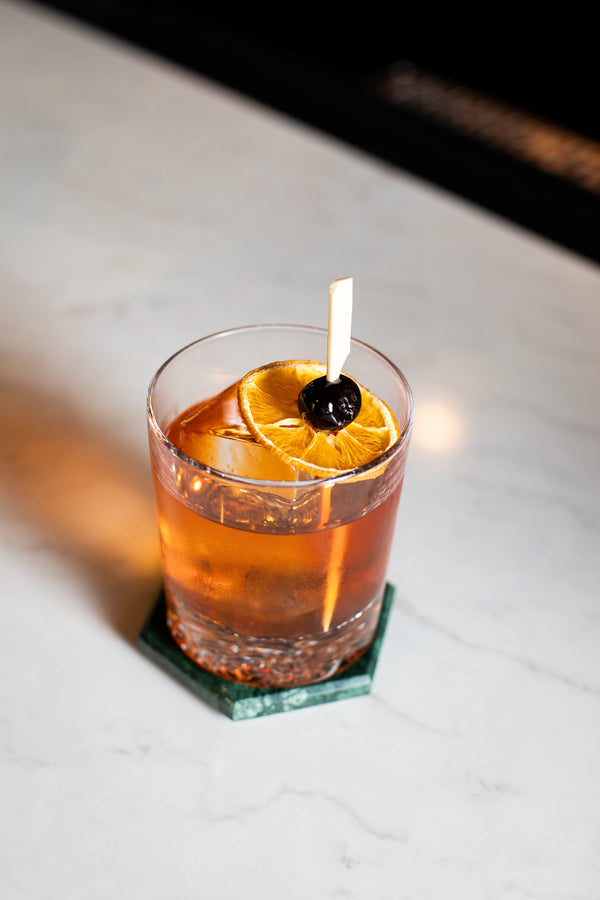 A deep amber cocktail in a clear rocks glass with a large ice cube, garnished with a dried orange wheel and dark cherry on a wooden pick, resting on a green marble hexagon coaster on a white marble surface