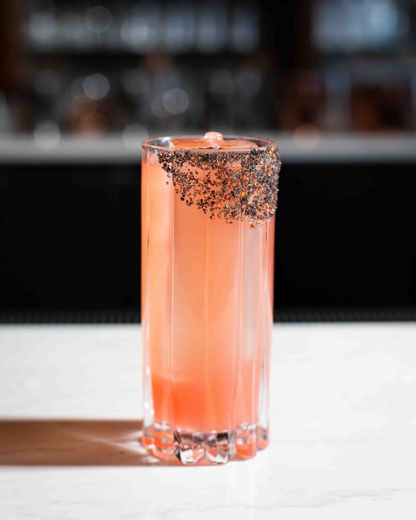 A tall coral pink cocktail in a ribbed highball glass with a bold black and red spiced rim, photographed against a dark bar background