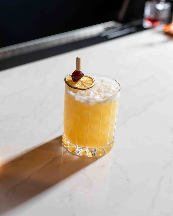 A golden yellow cocktail in a ribbed crystal rocks glass, topped with crushed ice and garnished with a dried lime wheel and a cherry on a pick, on a white marble surface