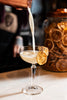 Cocktail being poured into a glass with a lemon wheel garnish, with a jar of dried fruit in the background.