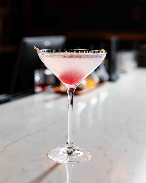 A pale white cocktail with a deep pink blush cherry resting at the base, served in a classic triangle martini glass on a white marble bar