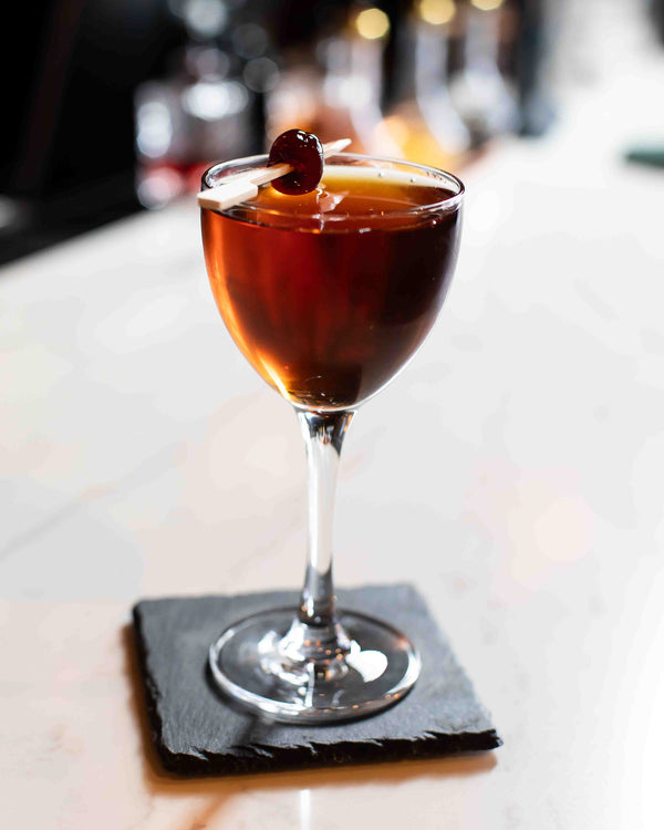A deep mahogany cocktail in a stemmed wine-style glass garnished with a dark cherry and orange peel on a pick, resting on a black slate coaster on a white marble bar