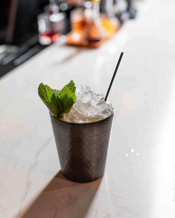 A cocktail in a dark hammered metal julep cup, filled with crushed ice and garnished with a large fresh mint bouquet and a thin black straw, on a white marble bar
