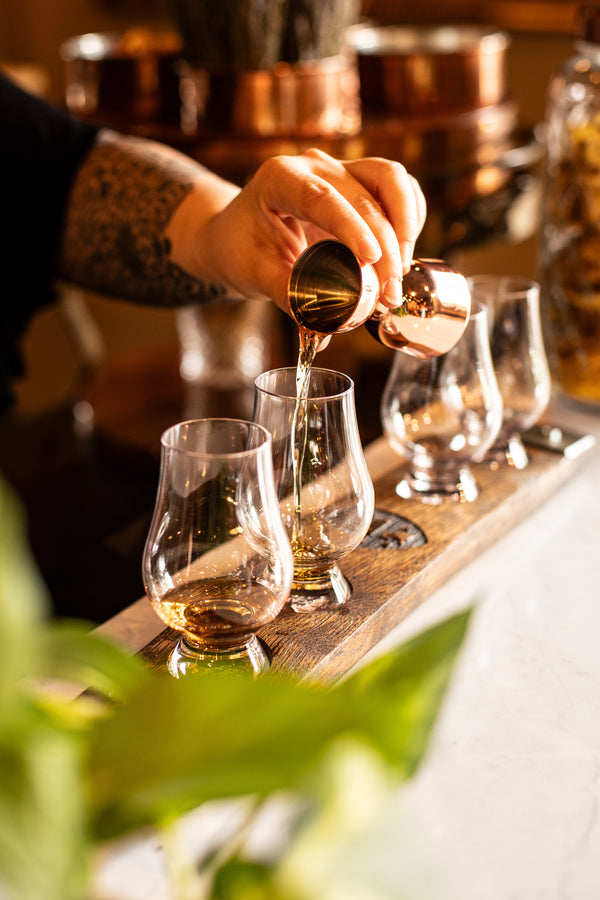 A hand pouring amber bourbon from a copper jigger into one of four Glencairn glasses lined up on a wooden flight board, copper pots visible in the background