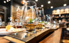 Glencairn whisky glasses with whisky on a wooden bar counter in a dimly lit bar.