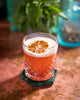 Glass of Negroni cocktail with foam and orange peel on a coaster, blurred plant and container in the background