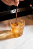 Glass of amber liquid with ice being poured on a marble surface