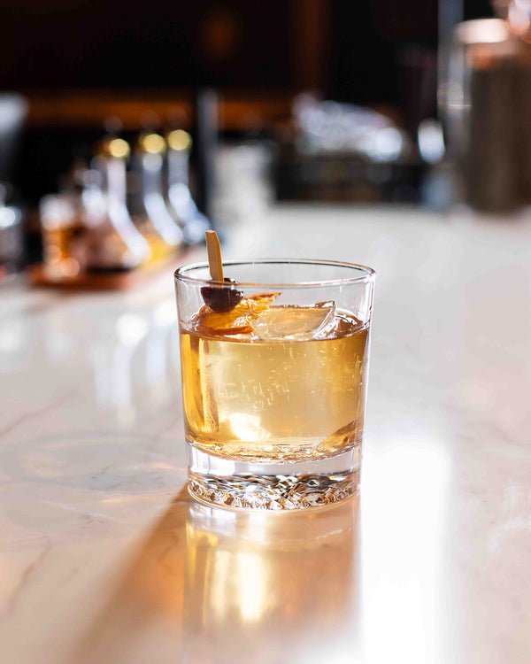 A golden amber cocktail in a clear rocks glass with a large ice cube, garnished with a dried cherry and orange slice on a wooden pick, on a white marble bar with a warm blurred background