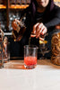 Person pouring a red cocktail into a glass with a bar setting in the background