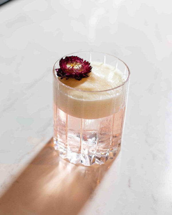 A pale pink cocktail in a clear fluted rocks glass, topped with a thick white passion fruit foam and a single dried burgundy flower, served on a white marble surface