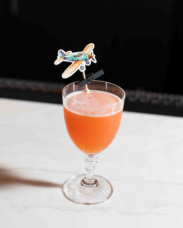 A bright orange cocktail in a stemmed goblet glass with a light foam top, garnished with a colorful vintage airplane cocktail pick, set against a dark background