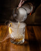 Smoked Manhattan Cocktail being poured into a glass with smoke and an orange peel garnish on a wooden surface.