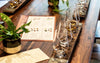 Wooden bar counter with glasses of whiskey, a menu, and a plant.