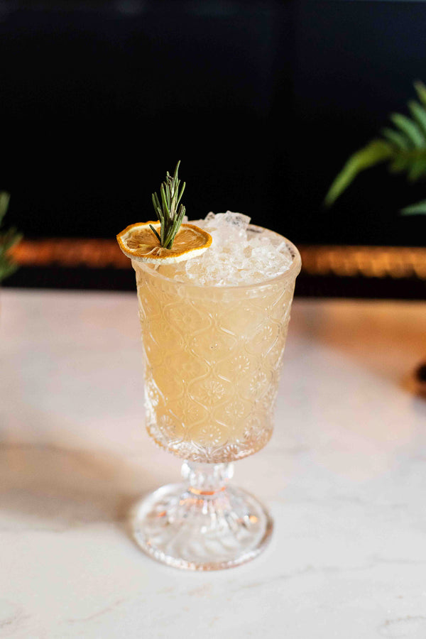 A golden yellow cocktail in an ornate crystal goblet filled with crushed ice, garnished with a fresh rosemary sprig and a dried orange wheel, set against a dark background with green ferns