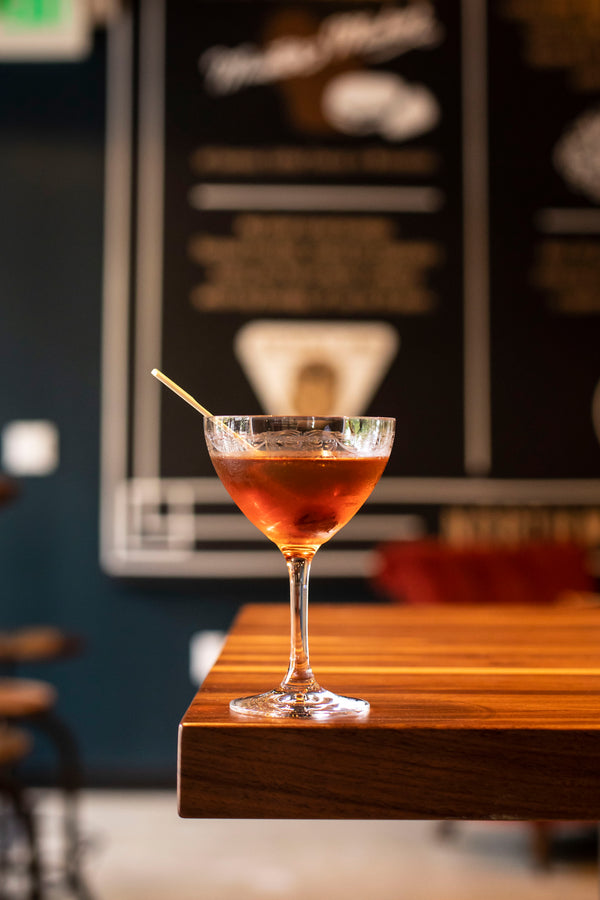 A deep amber cocktail in an etched crystal coupe glass with a bourbon cherry on a wooden pick, perched on the edge of a warm wood bar top with a dark menu board visible in the background