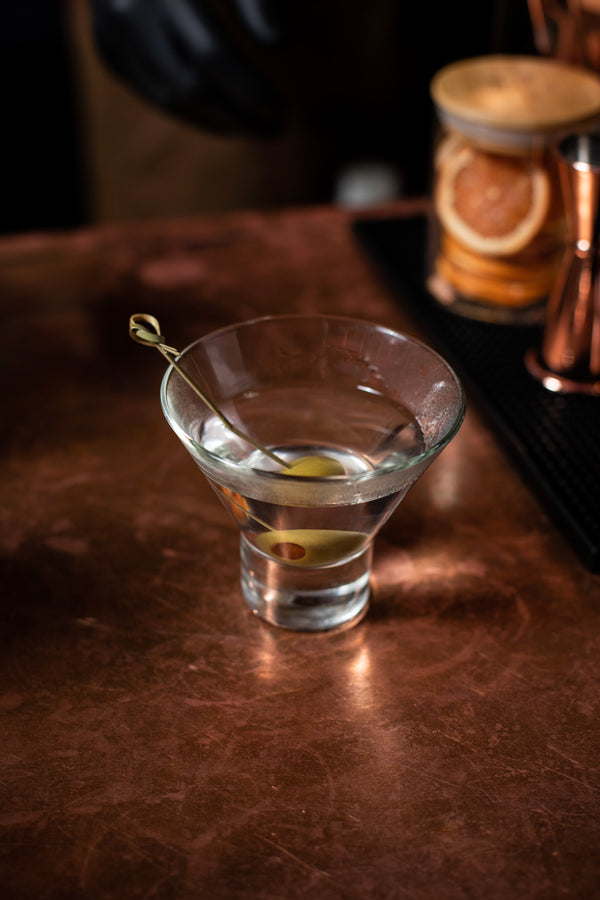 A classic martini in a chilled Nick and Nora glass with a single olive on a bamboo pick, resting on a warm copper bar top