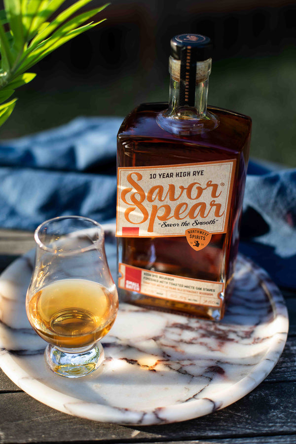A square amber glass bottle of Savor Spear 10 Year High Rye bourbon beside a small Glencairn glass of poured whiskey, resting on a white and red marble tray outdoors