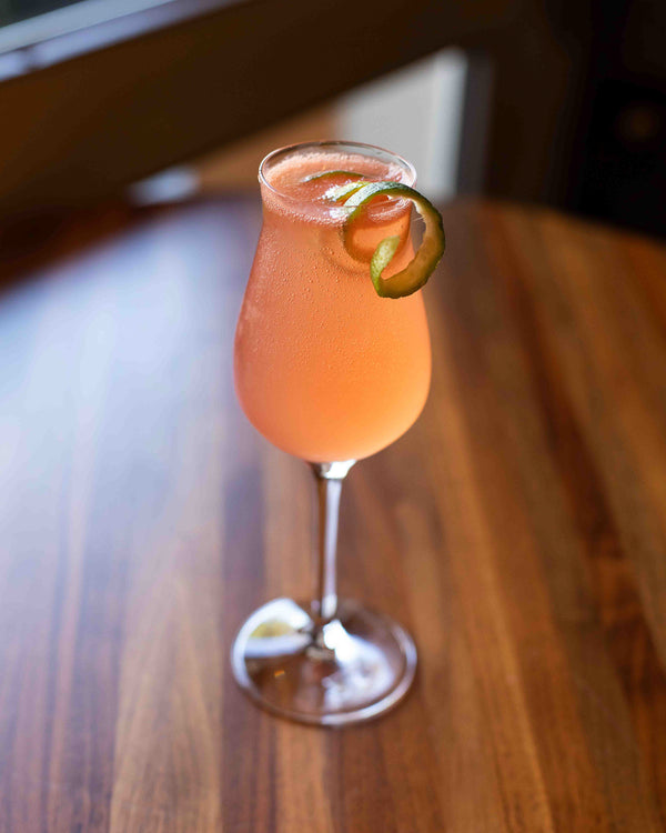 A bright coral pink cocktail in a tulip stem glass, garnished with a lime twist, set on a warm wood surface