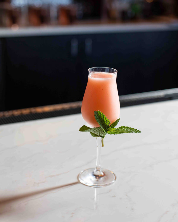 A peachy pink cocktail in a tulip stem glass with a large fresh mint bouquet tucked at the base of the glass, photographed on a white marble surface