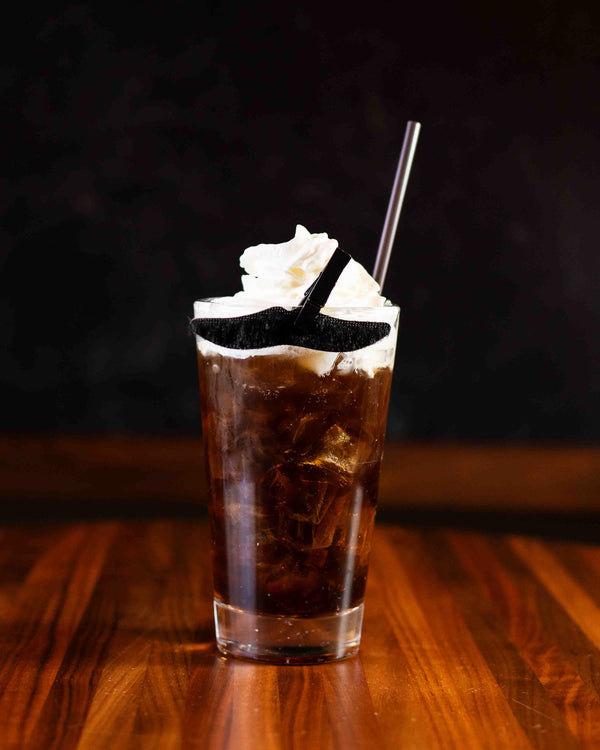 A dark iced cocktail in a pint glass topped with a generous swirl of whipped cream and a black wafer garnish, with a metal straw, on a warm wood surface against a dark background