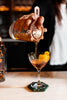 Bartender pouring a cocktail into a glass with a lemon twist, using a copper strainer.