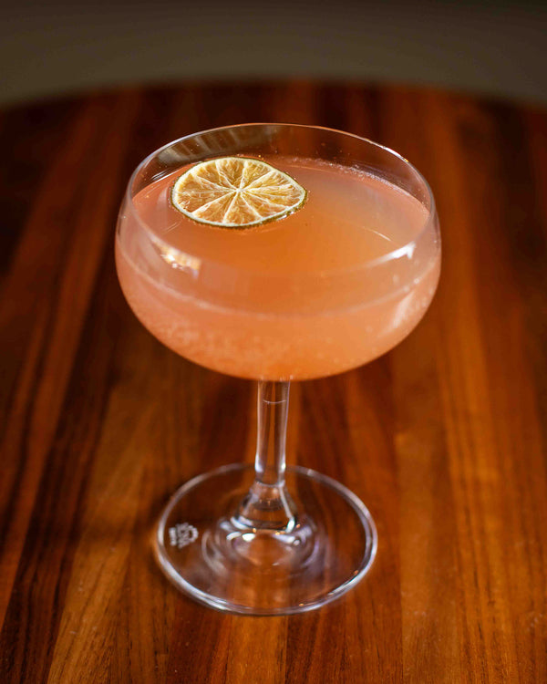 A soft pink cocktail in a wide coupe glass, garnished with a single dried lime wheel floating on the surface, photographed on a warm wood bar top