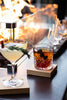 Glass of dark liquid on a coaster with a blurred fire pit in the background